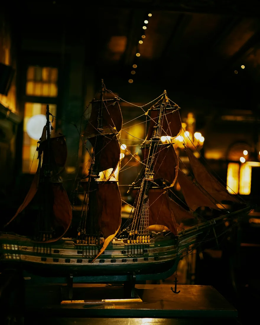 Modelo de barco de tres palos con jarcia y velas oscuras iluminado lateralmente sobre su peana